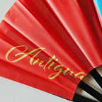 Close up of Antigua Barbuda flag inspired bamboo folding hand fan displaying the country's name in script font and displaying the Caribae Kiss logo.