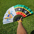 Handheld bamboo folding hand fan with inspirational design of the Dual Heritage Fan - US Virgin Islands + St. Kitts-Nevis flags colorful design held against a grassy background