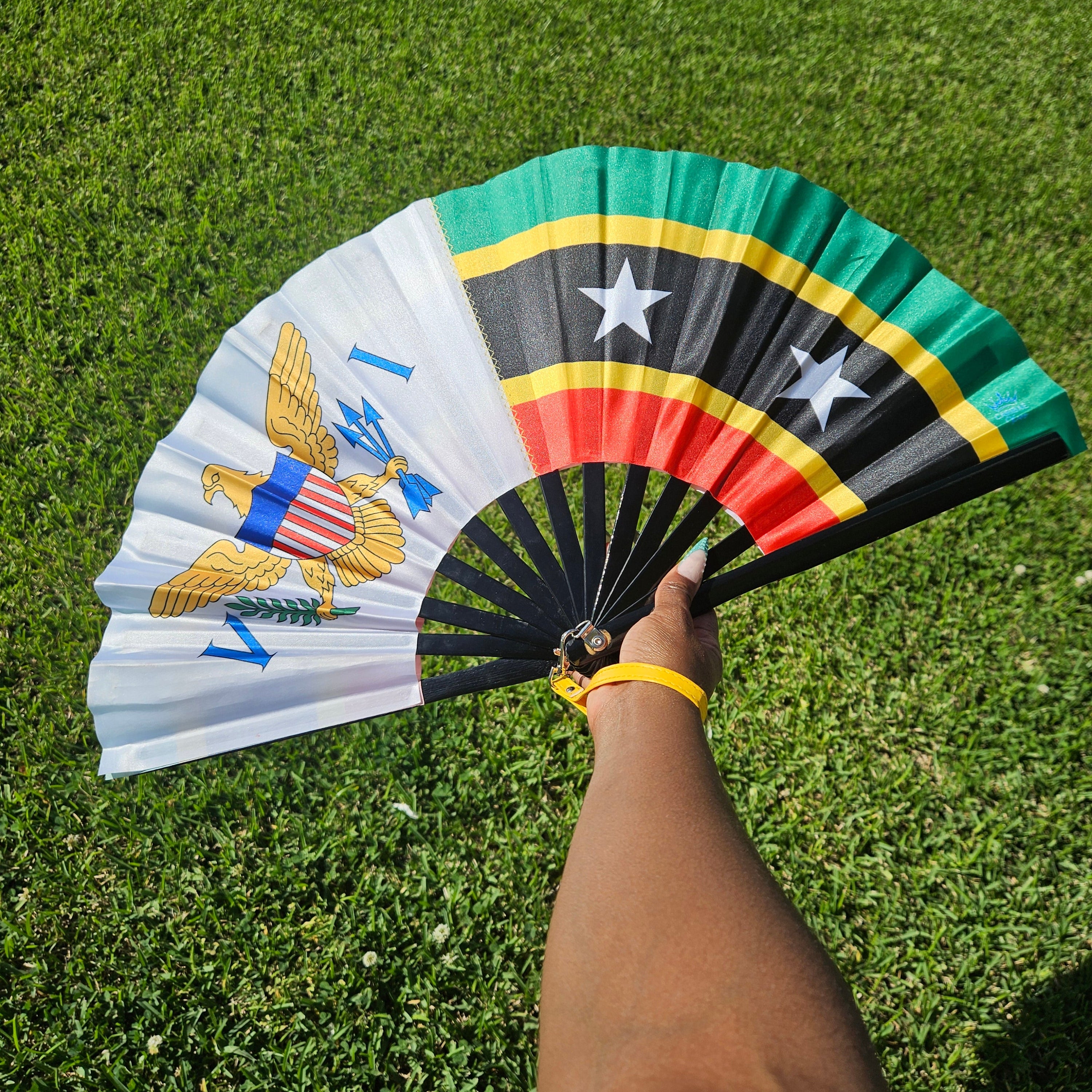 Handheld bamboo folding hand fan with inspirational design of the Dual Heritage Fan - US Virgin Islands + St. Kitts-Nevis flags colorful design held against a grassy background