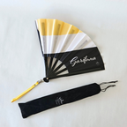 Garifuna flag inspired bamboo folding hand fan halfway open along with black suede satchel bag with Caribae Kiss white logo print.