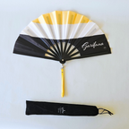 Flat lay of Garifuna flag inspired bamboo folding hand fan along with black suede satchel bag with Caribae Kiss white logo print.
