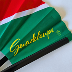 Close up of Guadeloupe flag inspired bamboo folding hand fan displaying the country's name in script font and displaying the Caribae Kiss logo.
