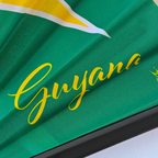 Close up of Guyana flag inspired bamboo folding hand fan displaying the country's name in script font and displaying the Caribae Kiss log