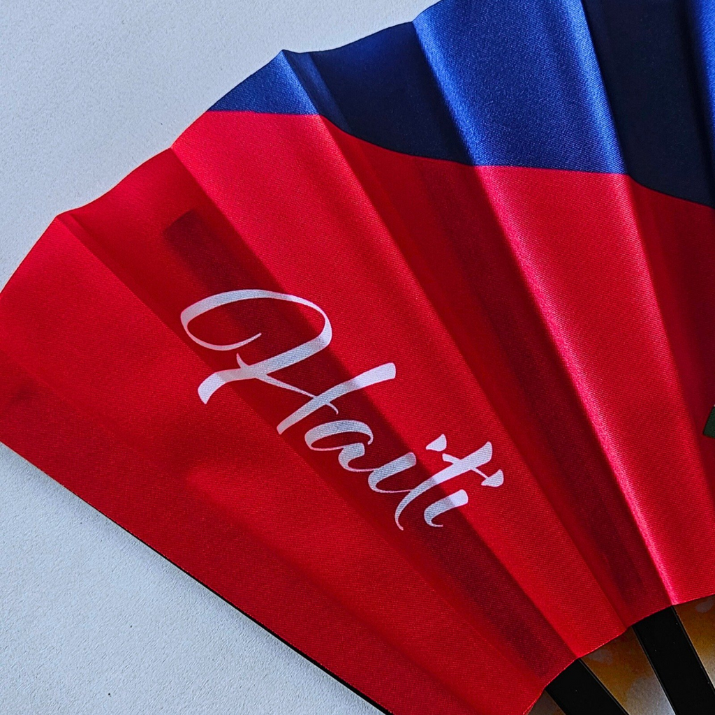 Close up of Haiti "Ayiti" flag inspired bamboo folding hand fan displaying the country's name in script font and displaying the Caribae Kiss logo.