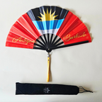 Flat lay of Antigua and Barbuda flag inspired bamboo folding hand fan along with black suede satchel bag with Caribae Kiss white logo print.